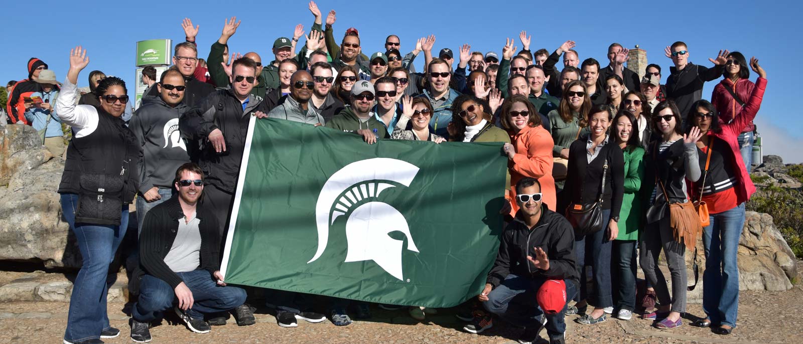 Large group of Michigan State Executive MBA students stand with MSU Spartan Helmet flag on sand in South Africa