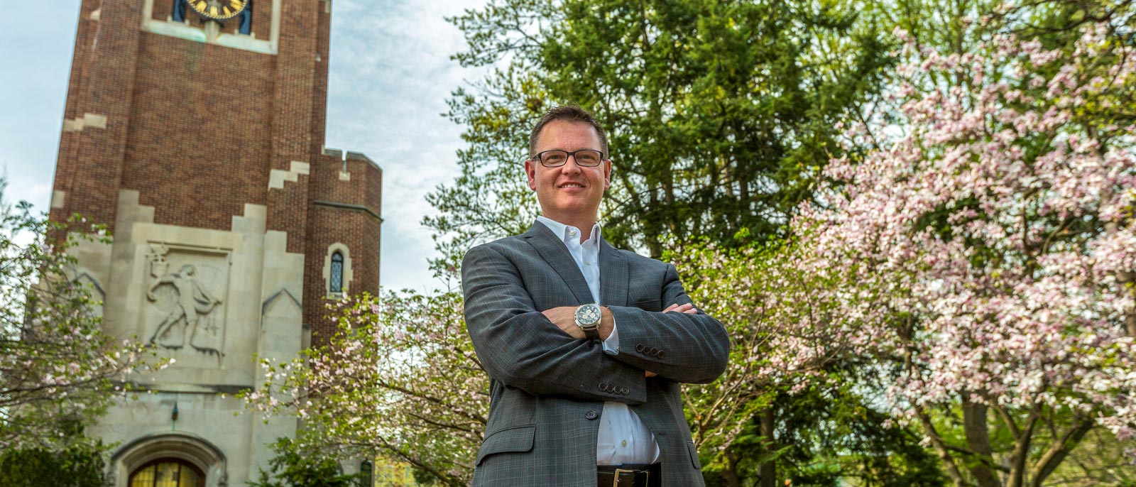 M.S. MSL graduate Jeff Day stands with arms crossed in front of Beaumont Tower and blooming flowering trees on the campus of MSU