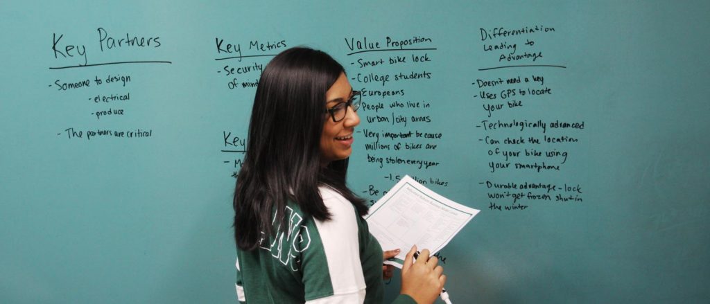 Female student turns around and smiles while writing business model details on a dry erase board.