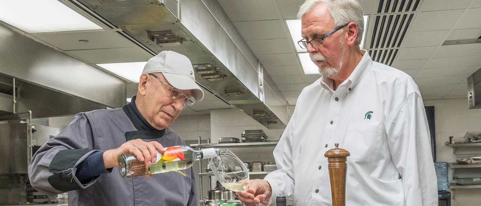Michigan State University professor Allan Sherwin pours a glass of wine for former The School of Hospitality Business Director Carl Borchgrevink, standing inside a commercial kitchen.