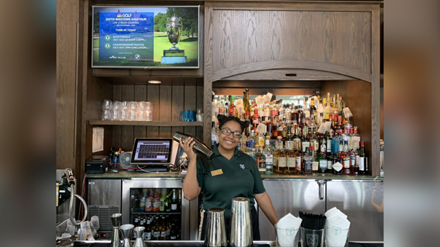 Foressia Hood stands behind a well-stocked bar, holding a drink shaker and smiling