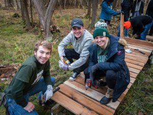 Three Spartans work together to build a bridge in a wooded area.
