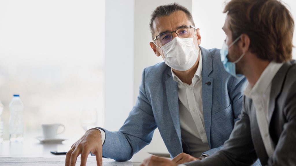 Businessmen sitting at conference table discussing business, wearing protective face mask due to COVID-19 pandemic