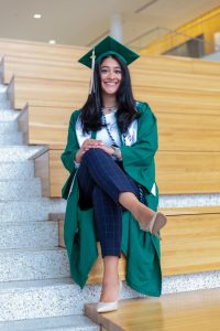 Sachi Arora sits on the grand staircase in the Minskoff Pavilion wearing her graduation cap and gown.