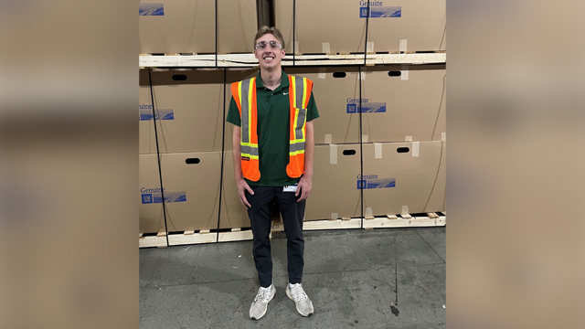 Alec Woodman in front of boxes on shelves wearing an Orange Safety Vest