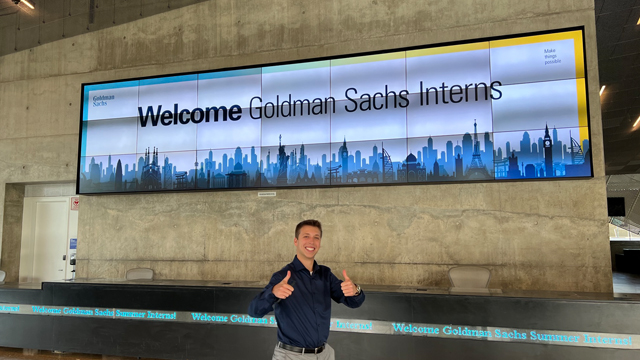 Blake Maday posing to camera with both thumbs up in front of the "Welcome to Goldman Sachs Summer Interns" Sign