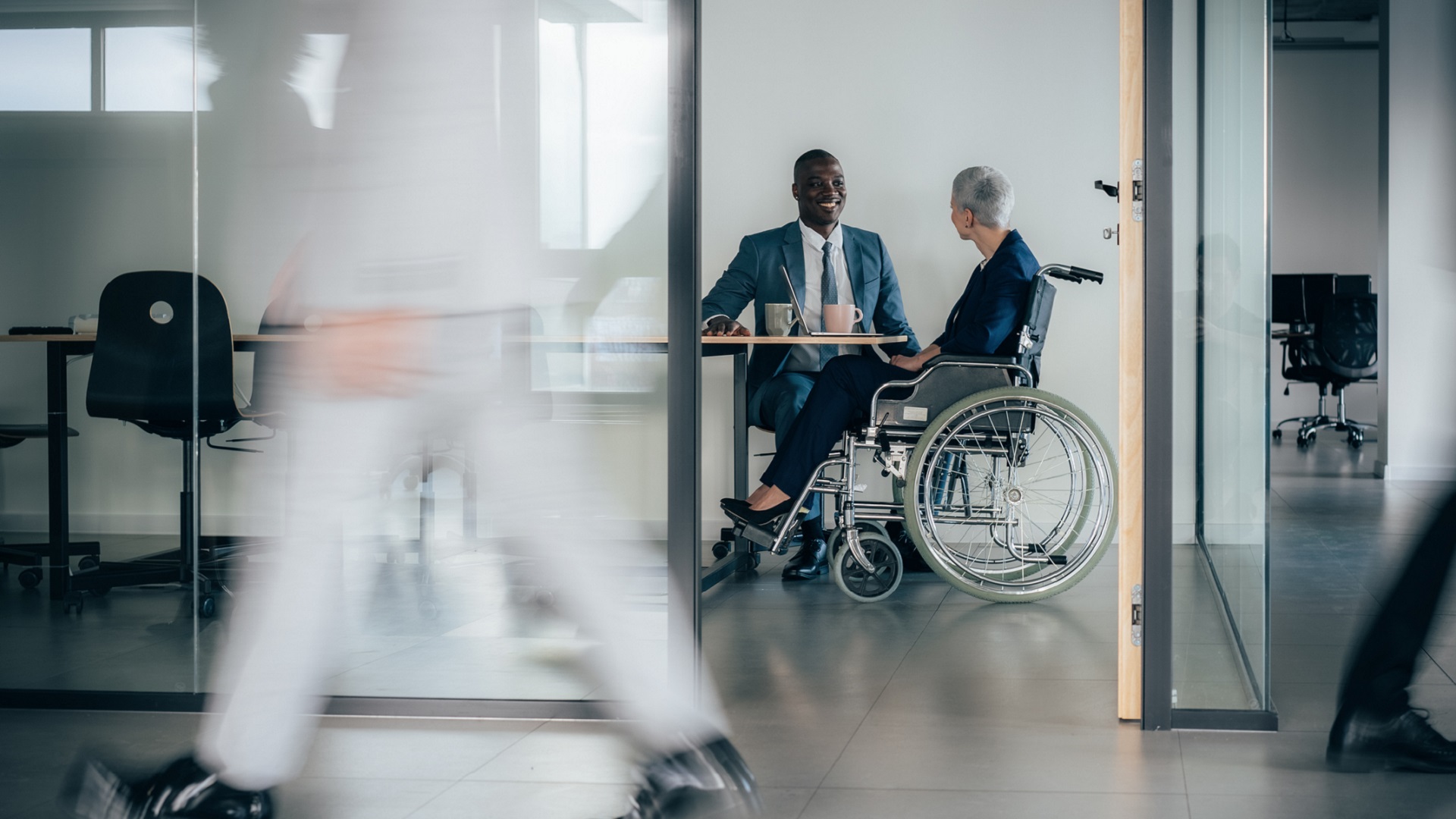 Business woman in wheelchair having meeting in office, someone walks by in the foreground blurred motion