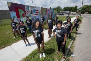 Members of Michigan nonprofit Detroit Heals Detroit wear matching tshirts stand on a sidewalk in front of a painted mural in Detroit.