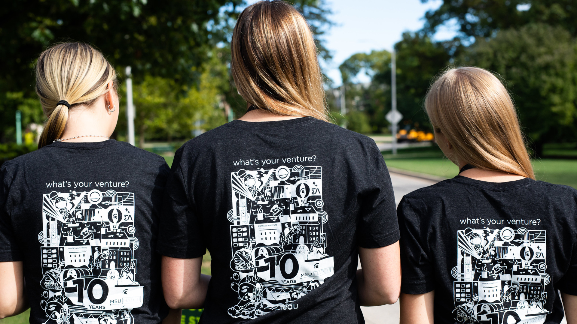 Three female students wearing matching entrepreneurship minor t-shirts with their backs to the camera outside on MSU's campus in summer.