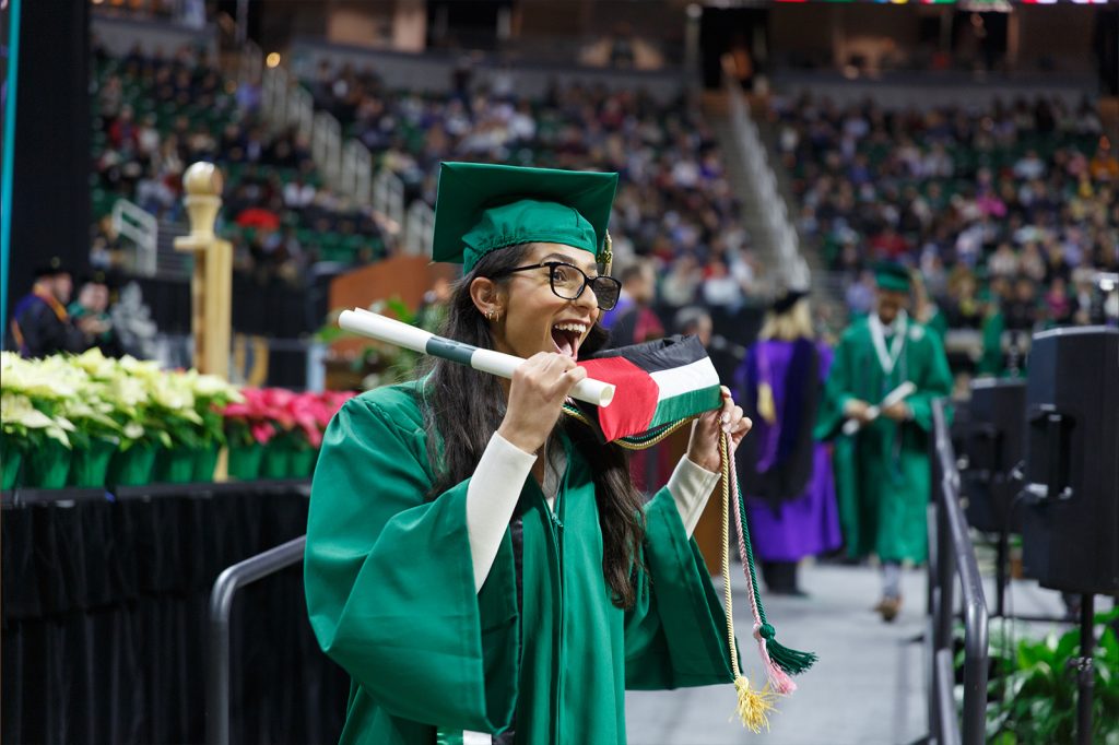 A Broad undergraduate student smiles while crossing the stage at commencemnt, holding her diploma and a Palestine flag.