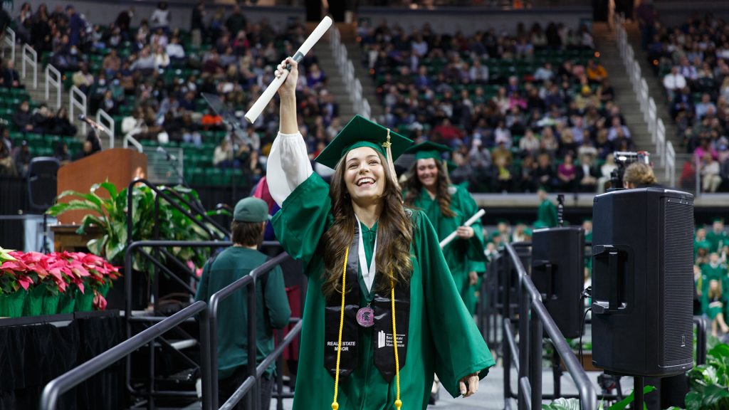 An undergrad student from the class of 2022 crosses the commencement stage, smiling while holding her diploma in the air.