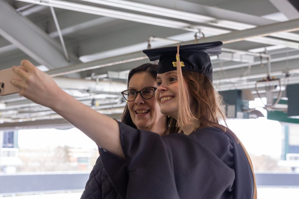 Woman MBA student wearing acadmeic regalia takes a selfie with her mom during commencement.