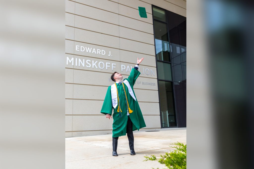 Carlos Conrad wears his graduation gown and tosses his cap into the air in front of the Minskoff Pavilion's main entrance.
