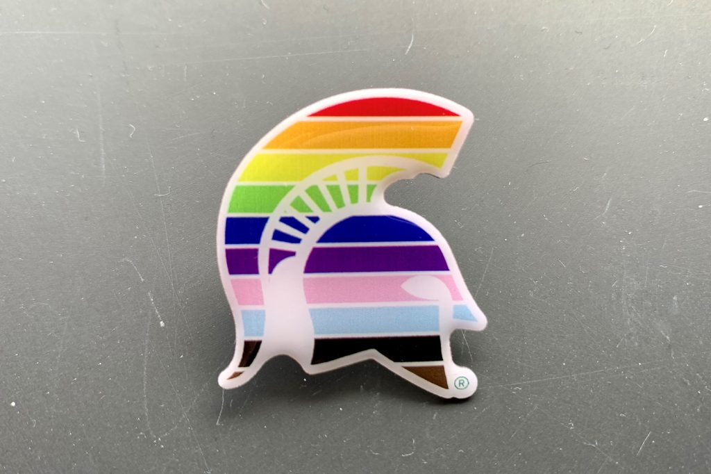A Spartan helmet lapel pin colored in LGBTQ+ pride stripes.
