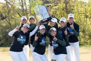 Women's golf team members wear Big 10 Championship t-shirts and hoist a trophy up in celebration.