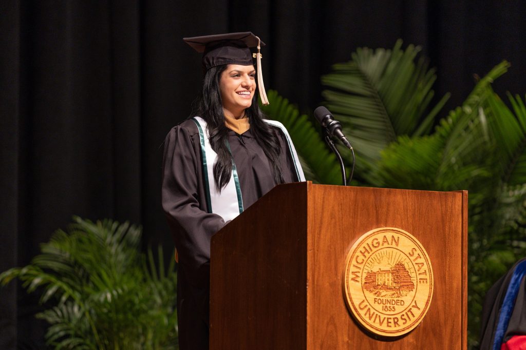 Prenella Semma dressed in academic regalia speaking from the podium on stage at the Wharton Center for the Class of 2023 EMBA graduation ceremony.