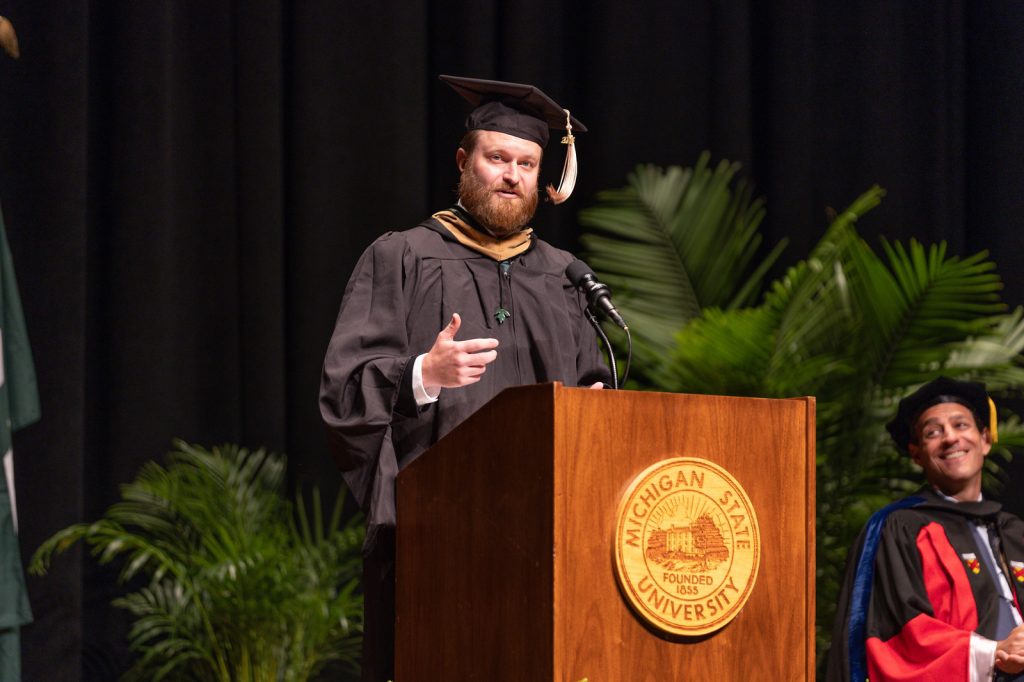 Tyler Wheeler dressed in academic regalia speaking from the podium on stage at the Wharton Center for the Class of 2023 EMBA graduation ceremony.
