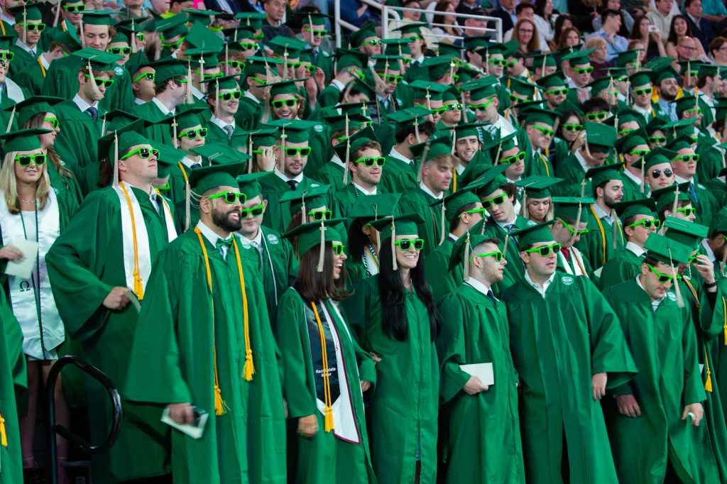 Busines undergraduate students fill the Breslin Center bleacher during commencement, wearing caps, gowns and neon green sunglasses.