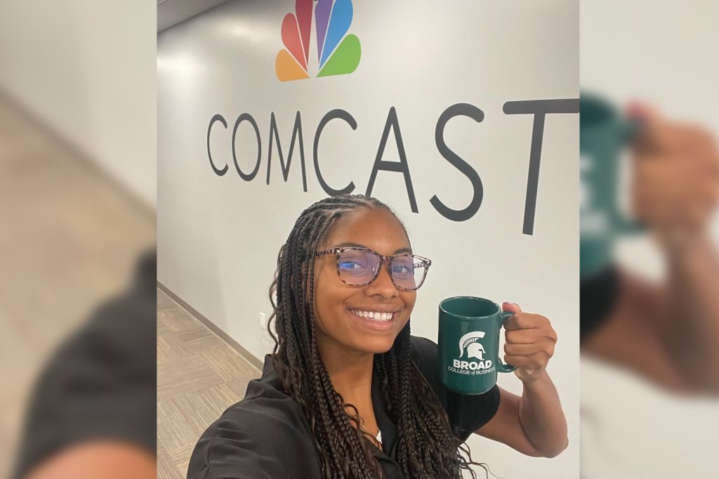 Symone King holds a Broad College of Business green coffee mug in front of a Comcast mural inside an office building during her 2023 internship experience.