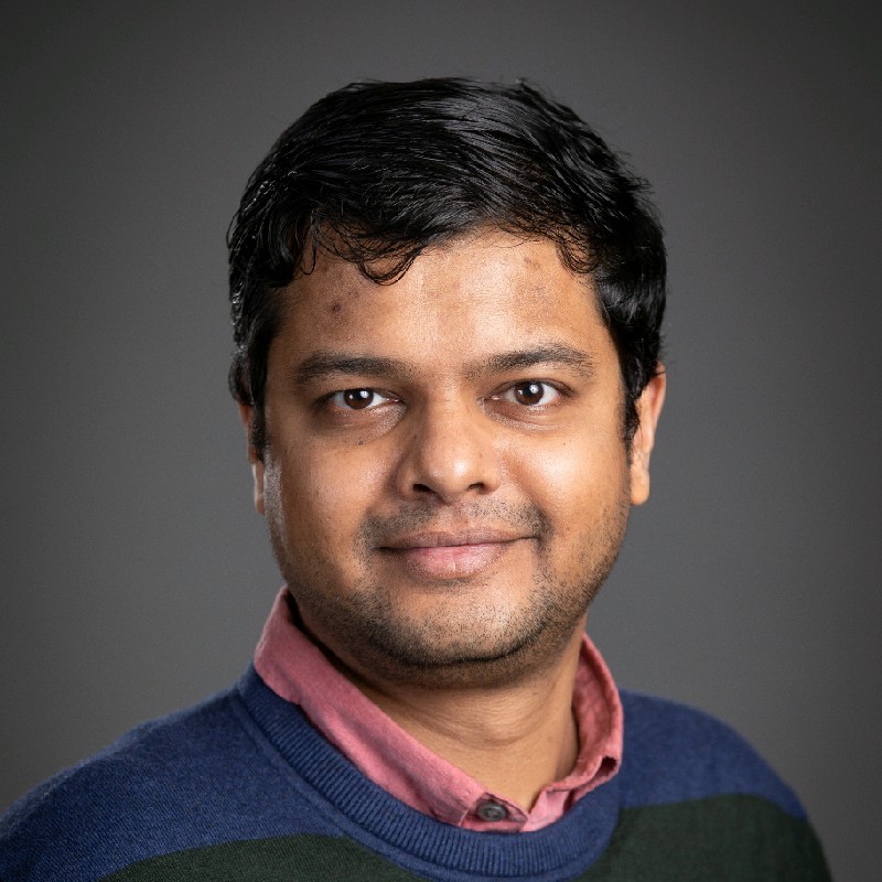 Headshot of man wearing a blue sweater with a pink under shirt.