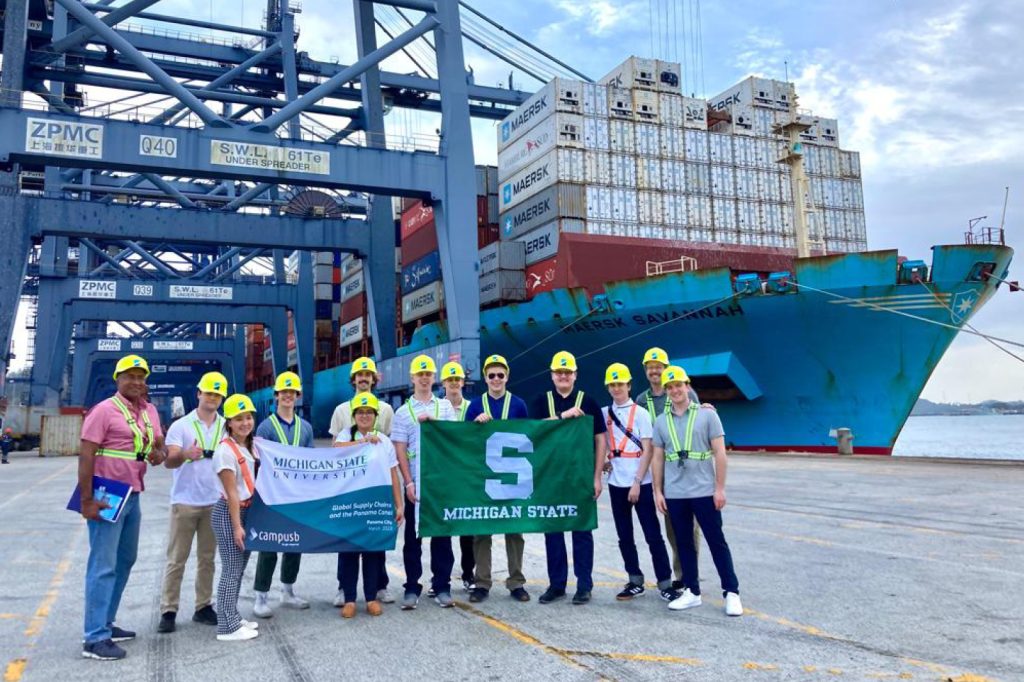 Students and staff pose for a picture with a Spartan flag near shipping freighters in Panama.