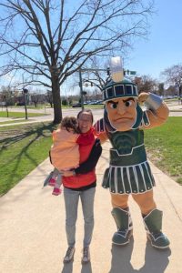Yuxuan Gao holding a child and posing with Sparty outside on a sunny day