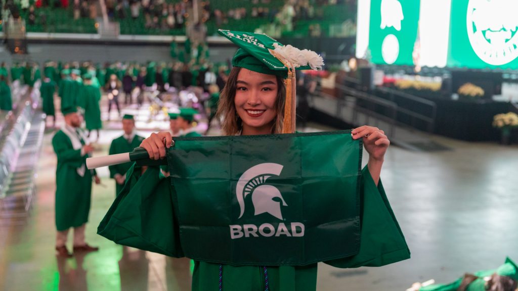 A Broad Spartan dressed in cap and gown, holds a Broad flag inside Breslin Center during MSU Commencement 2023.