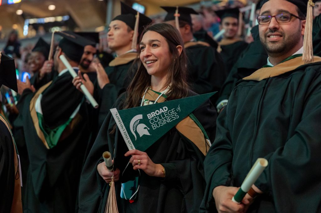 A student holds a Broad pennant and smiles in a crowd of peers at MSU's advanced degree commencement ceremony.