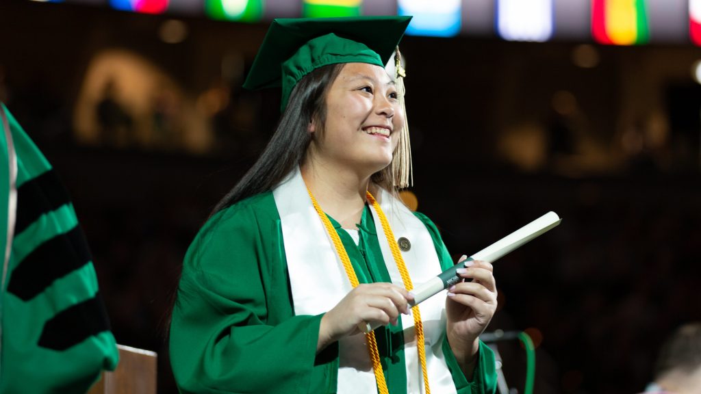 A Spartan holds her diploma and smiles while crossing the Breslin Center commencement stage.