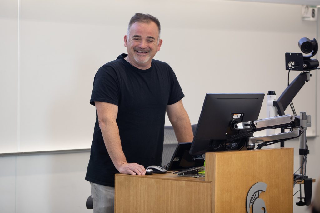 Dan Raynak leans on a podium and smilles while presenting to a Broad classroom.