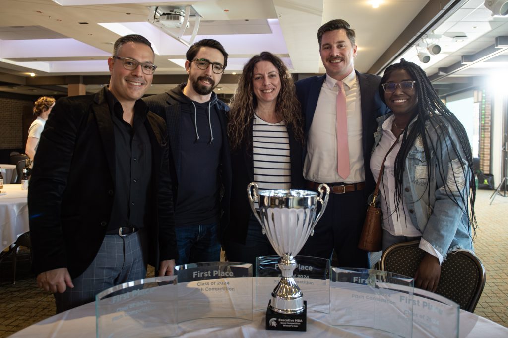 The 2024 EMBA Pitch Competition winning team stands together behind a table of trophies and awards.