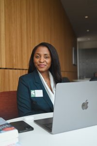 photographic a a black woman with long black hair in business attire with a laptop computer open on a desk in the foreground.