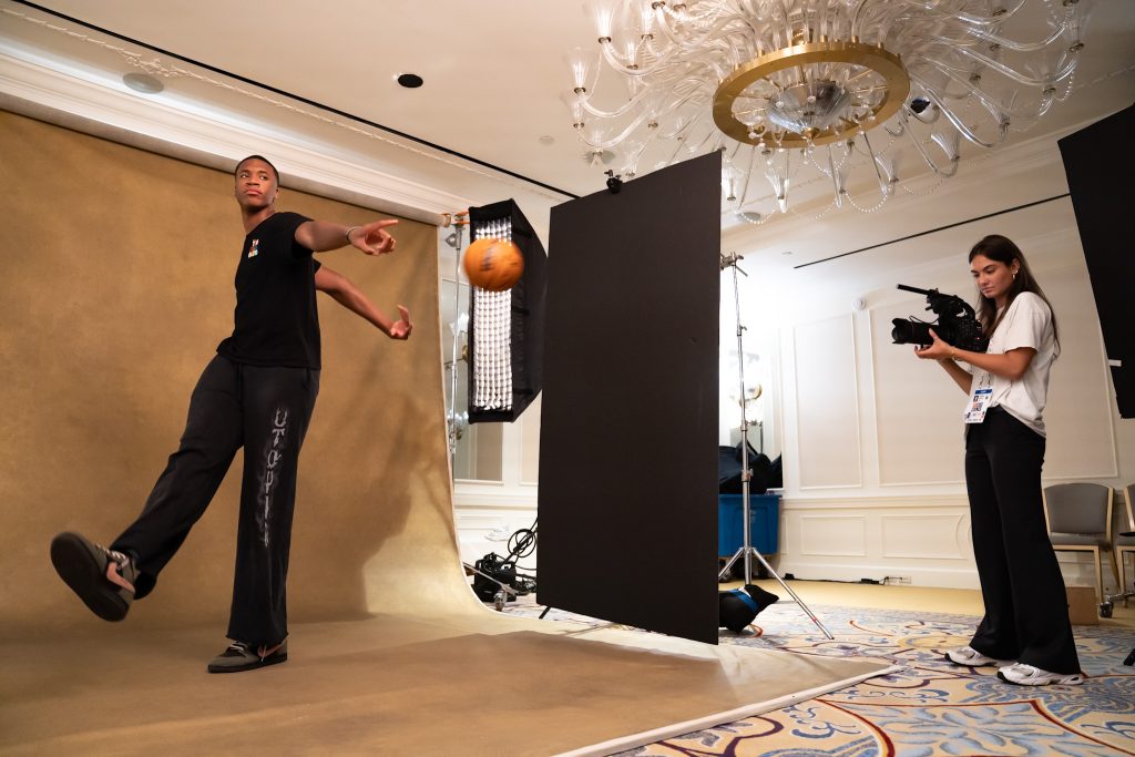 A basketball player poses with a basketball while a person with a camera films.