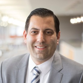 Headshot of man smiling wearing a grey jacket and blue/white tie against an office background.