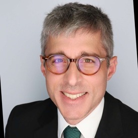 Headshot of man smiling wearing glasses and a black jacket against a grey background.