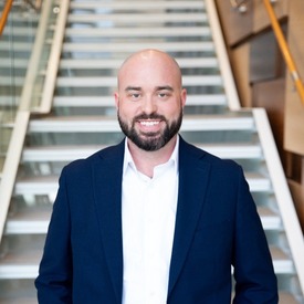 Headshot of man smiling wearing a blue jacket and white shirt against stairs.