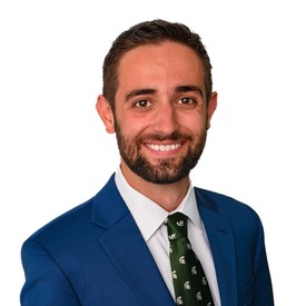 Headshot of man smiling with a blue jacket and green Michigan State tie. No background.