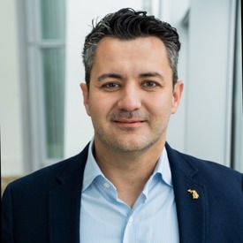 Headshot of man smiling wearing a dark blue jacket and light blue shirt against an office background.