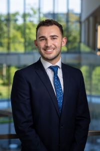 Headshot of Brandon Page, a young man with dark hair wearing a dark suit, white shirt, and blue tie