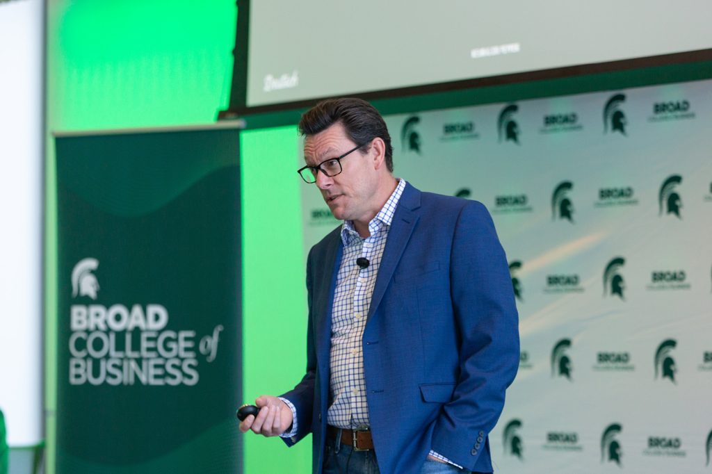 A speaker presents at the Broad College of Business, standing in front of a branded backdrop and holding a presentation remote.