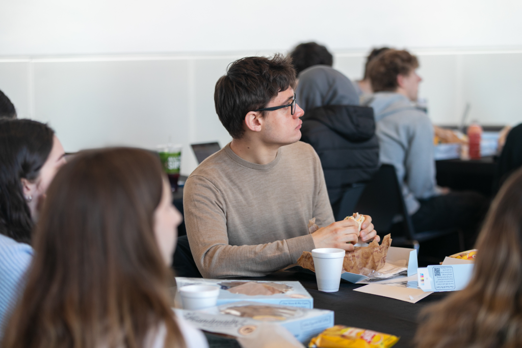 Students seated at round tables eat lunch and listen attentively during a campus event.
