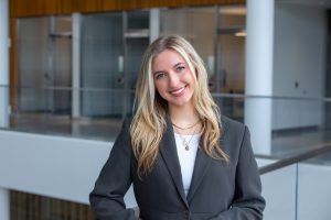 A smiling young woman with long blonde hair wears a gray blazer and layered necklaces, standing in a modern academic building with glass walls behind her.