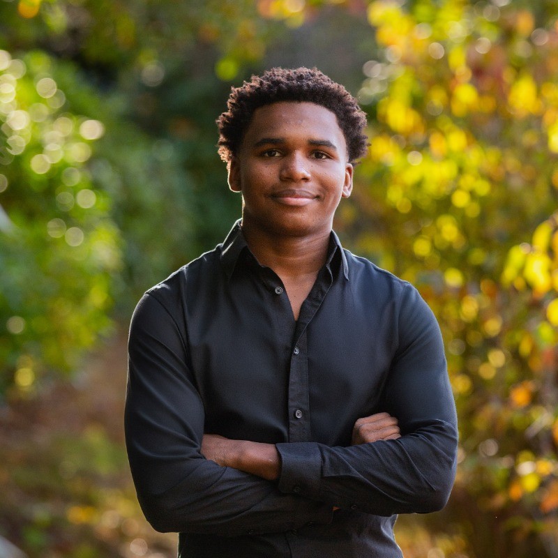 Young man in a black shirt standing in front of trees with his arms confidently crossed and smiling