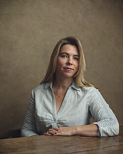 Sarah Wynn-Williams, framed in a chest-up portrait. Sarah has blonde and brown shoulder length hair and is wearing a white button down shirt with the sleeves rolled up.
