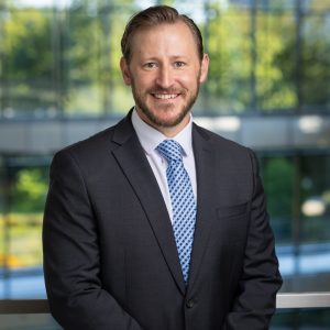 headshot of a white man with light brown hair and beard, wearing a business suit and a blue tie