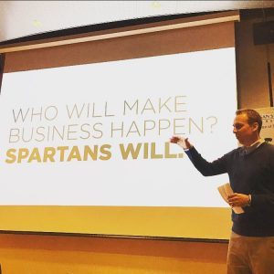 Ken Szymusiak next to his presentation. The slide reads "Who will make business happen? Spartans Will."