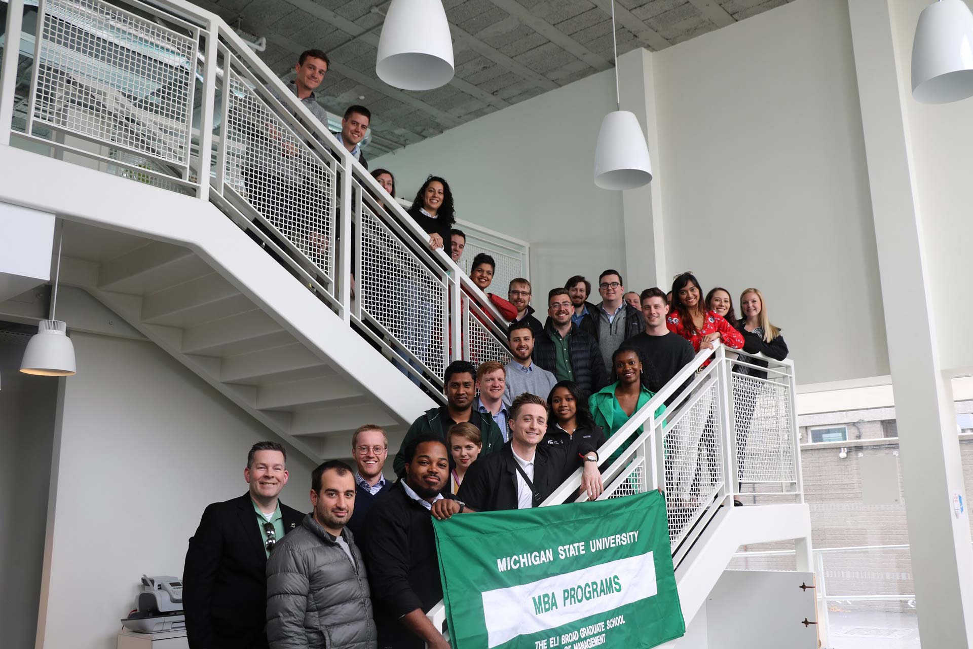 Group of full-time MBA students from the MSU Broad College of Business stand on stairs holding banner