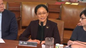 Ge Bai (Ph.D. Accounting and Information Systems ’12) dressed in a suit, seated and speaking into a microphone at a Congress hearing.
