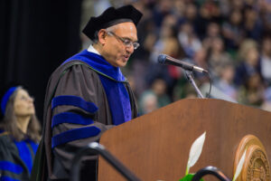 Dean Sanjay Gupta speaking at a podium for a graduation ceremony.