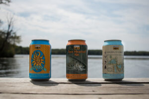 Bell's Brewery beer cans for Oberon, Two Hearted Ale and Lager of the Lakes on a wood surface in front of a body of water.